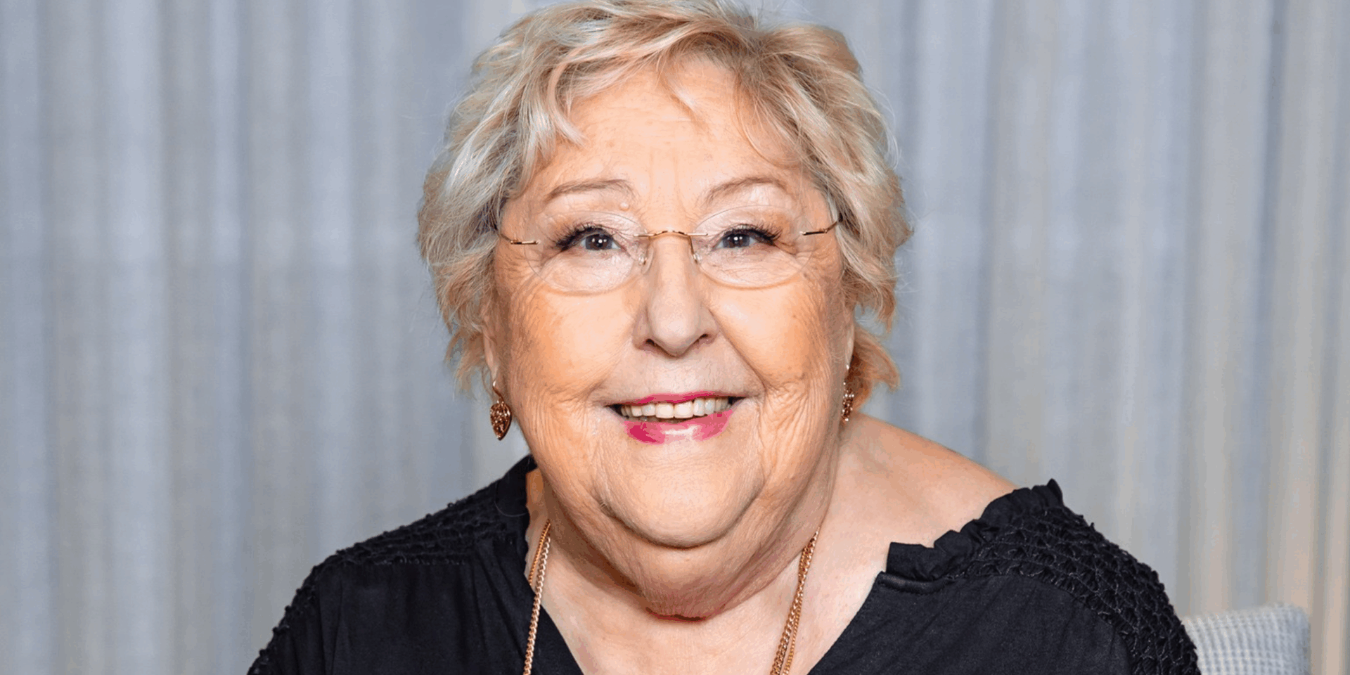 Photo of Sylvia Young with short blonde hair, glasses wearing a gold necklace and a black top in front of grey curtains.